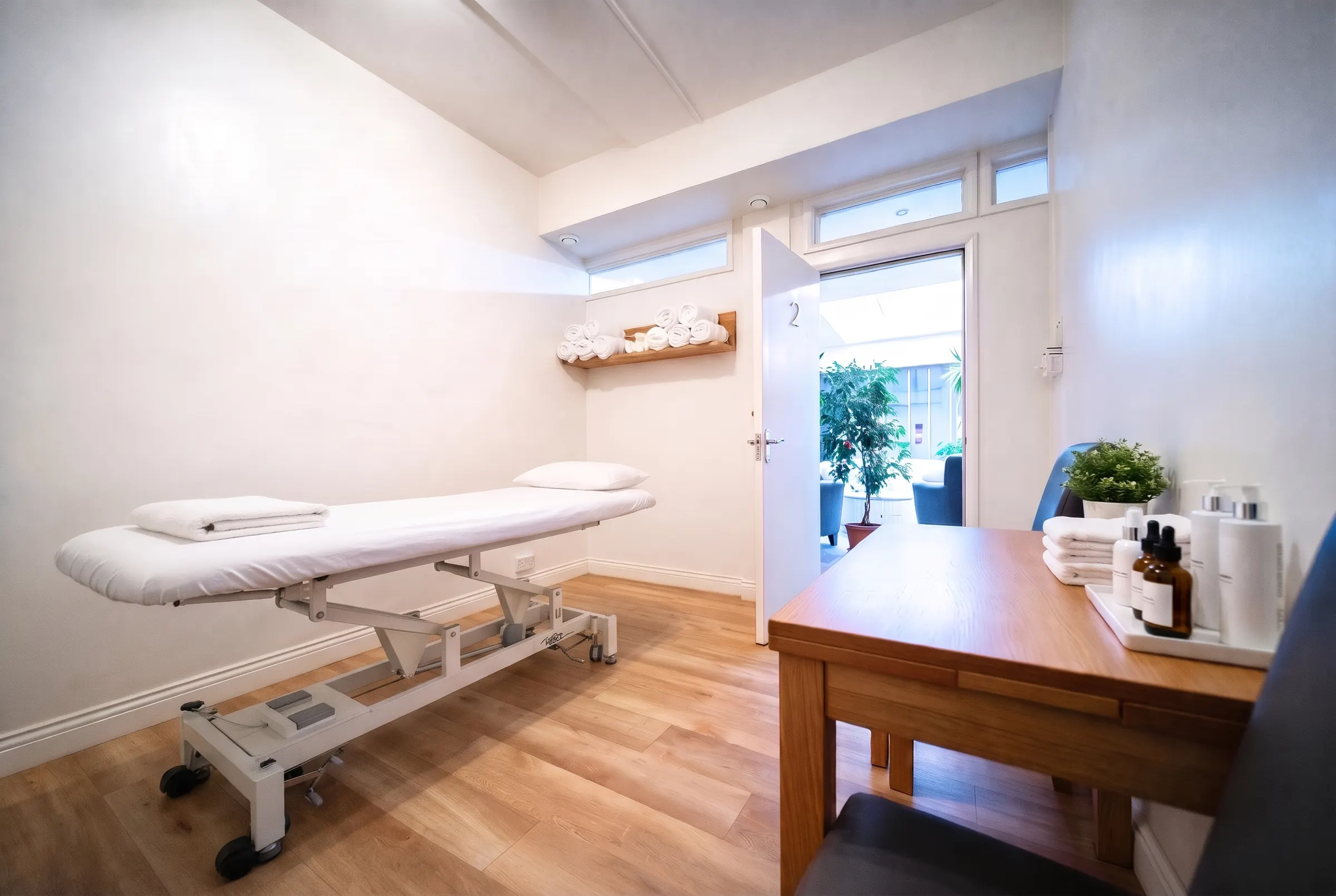 A treatment room at The Light Centre: a couch made up in white linen, rolled towels on a shelf and skincare products on the desk